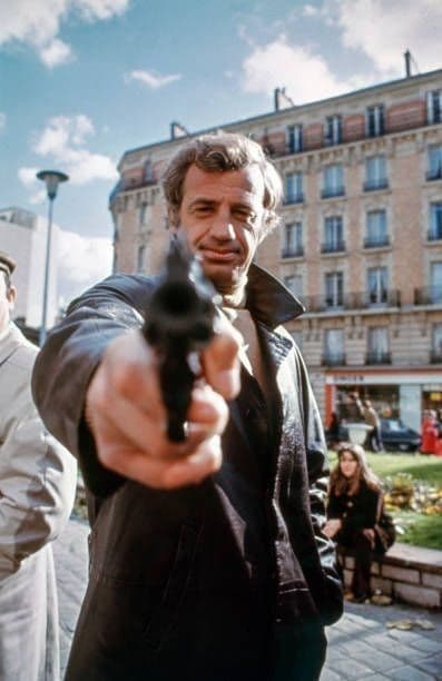 Jean-Paul Belmondo aiming a revolver straight at the camera on a Paris street