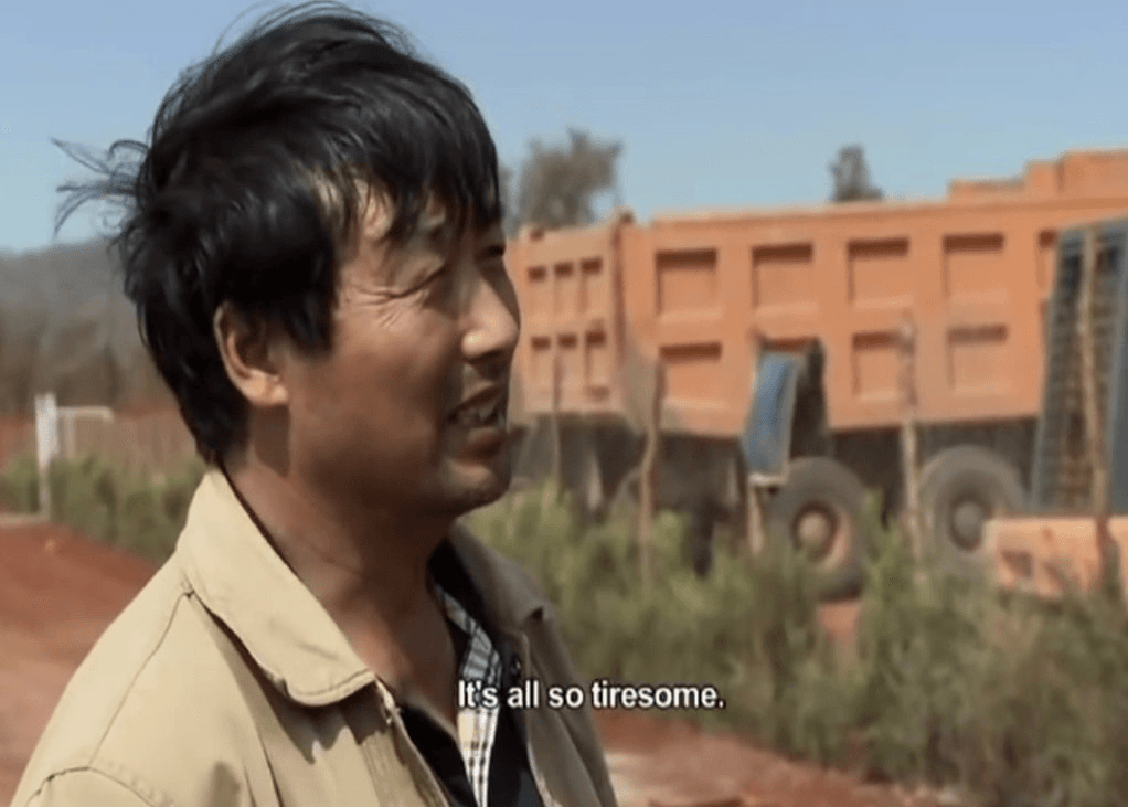 Man standing by a dirt road next to a dump truck, subtitled 'It's all so tiresome'